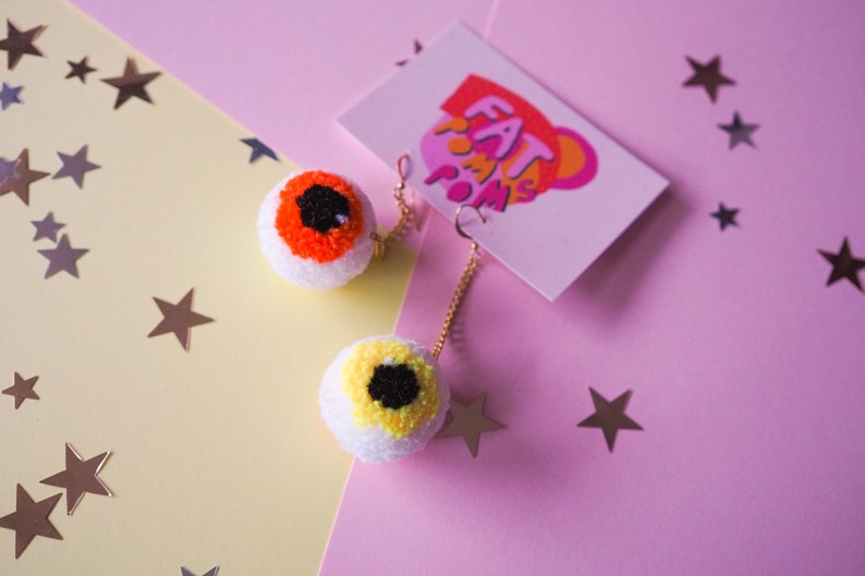 Puede incluir: Un par de pendientes de pompon en forma de ojo con cadenas doradas. Los pompones son blancos con detalles de hilo naranja y amarillo. Los pendientes est&aacute;n sobre un fondo rosa y amarillo con estrellas doradas.
