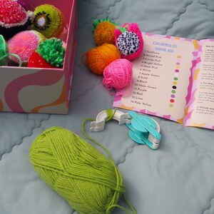 Puede incluir: Un kit de manualidades con una caja de pompones de colores con forma de frutas, como kiwi y fruta del drag&oacute;n. El kit incluye un ovillo de lana verde lima, un fabricante de pompones y una gu&iacute;a de colores con el texto "COLOURS IN YOUR KIT".