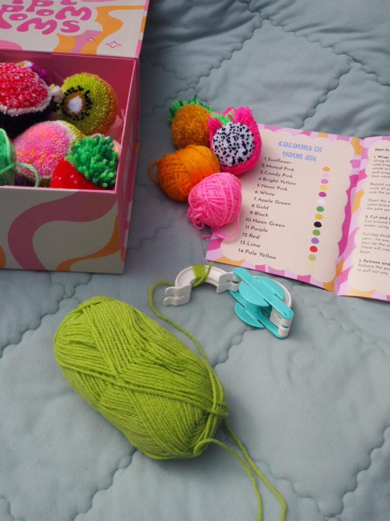 Puede incluir: Un kit de manualidades con una caja de pompones de colores con forma de frutas, como kiwi y fruta del drag&oacute;n. El kit incluye un ovillo de lana verde lima, un fabricante de pompones y una gu&iacute;a de colores con el texto "COLOURS IN YOUR KIT".