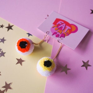 Puede incluir: Un par de pendientes de pomp&oacute;n blanco con ojos naranja y amarillo. Los pendientes cuelgan de cadenas doradas. Los pendientes est&aacute;n sobre un fondo rosa y amarillo con estrellas doradas.