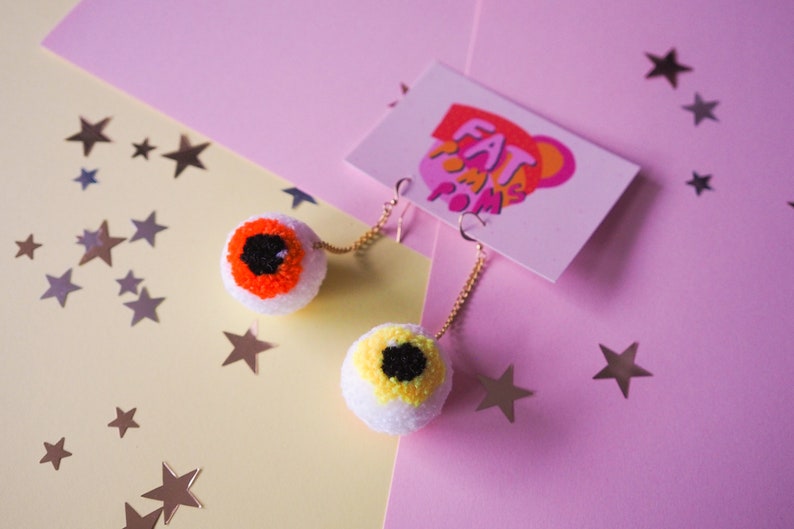Puede incluir: Un par de pendientes de pomp&oacute;n blanco con ojos naranja y amarillo. Los pendientes cuelgan de cadenas doradas. Los pendientes est&aacute;n sobre un fondo rosa y amarillo con estrellas doradas.