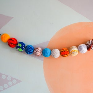 Puede incluir: Una colorida guirnalda de pompones de hilo en varios colores, incluyendo naranja, rojo, azul y blanco, ensartados en un hilo negro. Los pompones est&aacute;n dispuestos en una l&iacute;nea curva sobre un fondo pastel, creando un elemento decorativo y l&uacute;dico.