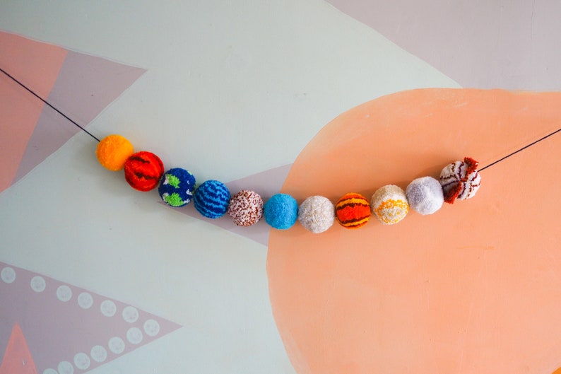 Puede incluir: Una colorida guirnalda de pompones de hilo en varios colores, incluyendo naranja, rojo, azul y blanco, ensartados en un hilo negro. Los pompones est&aacute;n dispuestos en una l&iacute;nea curva sobre un fondo pastel, creando un elemento decorativo y l&uacute;dico.