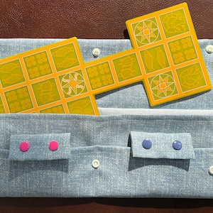 May include: A blue fabric pouch with two pockets and a yellow game board with fish and sun symbols. The pouch has two pink buttons and two blue buttons. The game board is made of cardboard and has a white background.