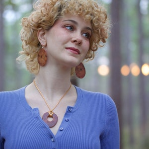 May include: A woman with curly blonde hair wearing a blue sweater and a wooden necklace with a circular pendant. She is also wearing wooden earrings.