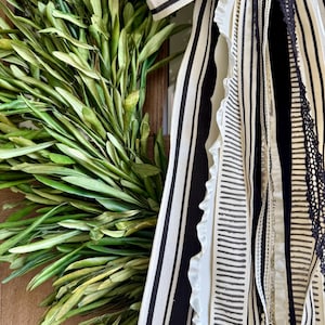 May include: A close-up of a decorative wreath featuring fresh green olive branches. The wreath is adorned with black and white striped ribbons and other decorative ribbons with various patterns and textures. The wreath is a natural home decor item.