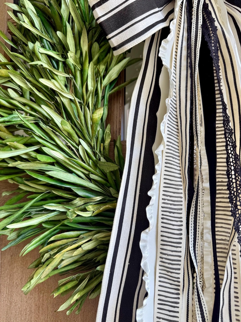 May include: A close-up of a decorative wreath featuring fresh green olive branches. The wreath is adorned with black and white striped ribbons and other decorative ribbons with various patterns and textures. The wreath is a natural home decor item.