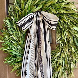 May include: A handmade wreath composed of green leaves, accented with a black and white striped bow and flowing ribbons. The ribbons include various patterns and textures, adding visual interest to the natural foliage. This decorative wreath is suitable for seasonal home decor.