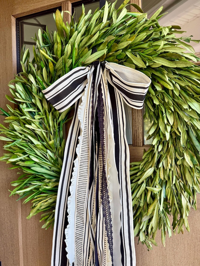 May include: A handmade wreath composed of green leaves, accented with a black and white striped bow and flowing ribbons. The ribbons include various patterns and textures, adding visual interest to the natural foliage. This decorative wreath is suitable for seasonal home decor.