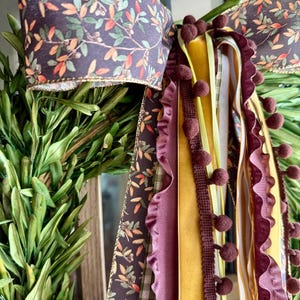 May include: A decorative bow with a dark purple fabric featuring a floral pattern. The bow has multiple ribbons in shades of yellow, purple, and green, some with pom-poms and ruffled edges. The bow is attached to a wreath of green leaves.