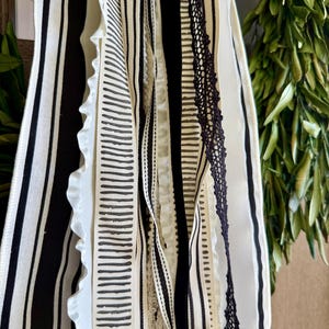 May include: A collection of decorative ribbons in various patterns and textures. The ribbons feature stripes, dotted lines, and lace in black and white. Some ribbons have ruffled edges, creating a layered effect. The background includes green foliage.