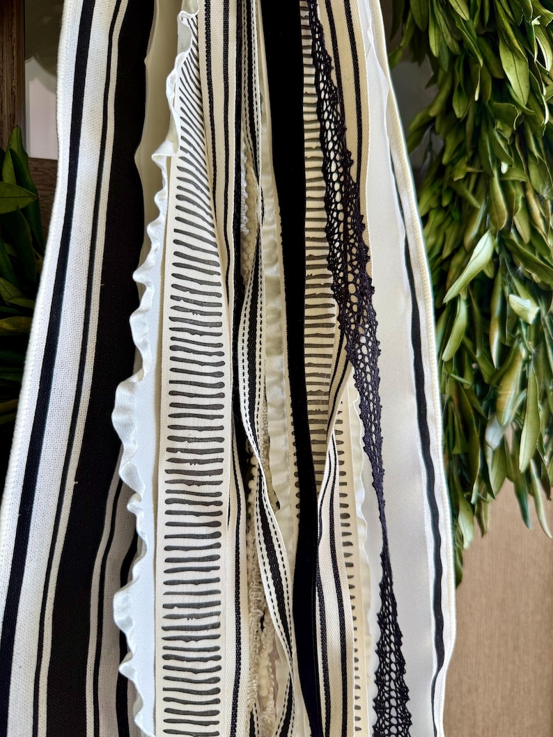 May include: A collection of decorative ribbons in various patterns and textures. The ribbons feature stripes, dotted lines, and lace in black and white. Some ribbons have ruffled edges, creating a layered effect. The background includes green foliage.