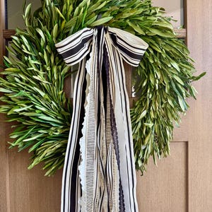 May include: A fresh green olive leaf wreath with a black and white striped bow and flowing ribbons. The wreath is hanging on a wooden door, creating a natural and elegant home decor accent.