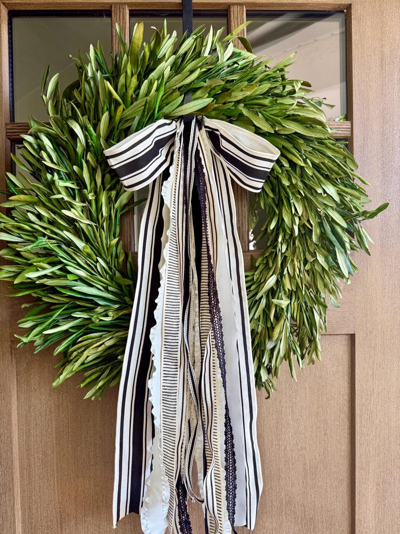 May include: A fresh green olive leaf wreath with a black and white striped bow and flowing ribbons. The wreath is hanging on a wooden door, creating a natural and elegant home decor accent.