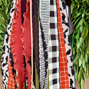Op de afbeelding: Close-up van een decoratieve krans met diverse linten. De linten hebben patronen zoals vleermuizen, ruitjes en strepen in zwart, oranje en wit. Sommige linten hebben gerimpelde randen en kanten details.