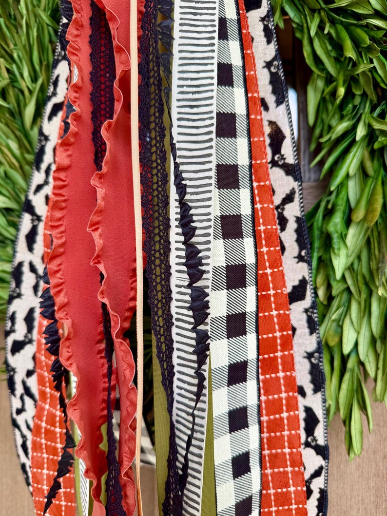 Op de afbeelding: Close-up van een decoratieve krans met diverse linten. De linten hebben patronen zoals vleermuizen, ruitjes en strepen in zwart, oranje en wit. Sommige linten hebben gerimpelde randen en kanten details.