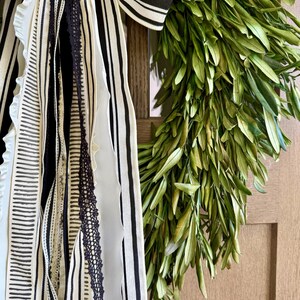 May include: A decorative wreath featuring a lush arrangement of green leaves. The wreath is adorned with a large bow and ribbons in black and white stripes and patterns, adding a touch of elegance.