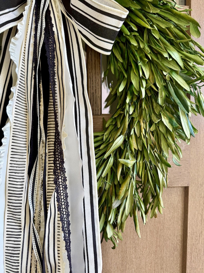 May include: A decorative wreath featuring a lush arrangement of green leaves. The wreath is adorned with a large bow and ribbons in black and white stripes and patterns, adding a touch of elegance.