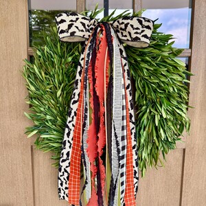 Op de afbeelding: Een groene krans met een decoratieve strik met een vleermuismotief. De strik is zwart en wit. De krans heeft oranje, zwarte en witte linten die naar beneden hangen. De krans hangt aan een houten deur.