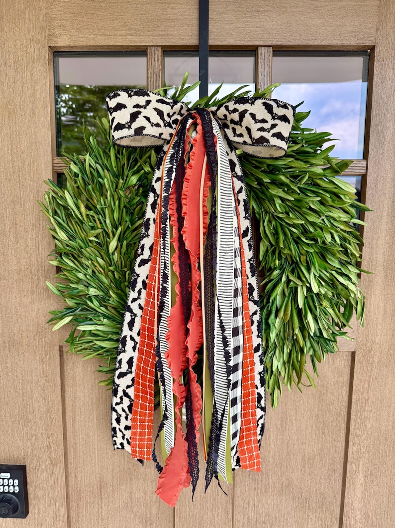 Op de afbeelding: Een groene krans met een decoratieve strik met een vleermuismotief. De strik is zwart en wit. De krans heeft oranje, zwarte en witte linten die naar beneden hangen. De krans hangt aan een houten deur.