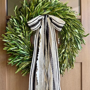 May include: A fresh olive leaf wreath with a black and white striped bow and cascading ribbons. The ribbons include various patterns and textures, adding visual interest to the natural green wreath. The wreath is hanging on a wooden door.
