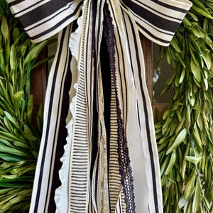 May include: A decorative wreath with a large black and white striped bow. The bow has multiple streamers in various patterns, including stripes and lace. The wreath is made of green leaves.