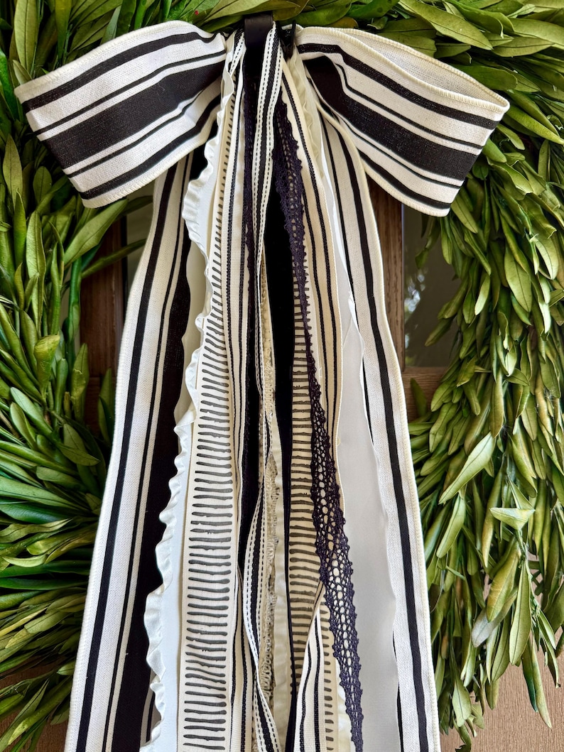 May include: A decorative wreath with a large black and white striped bow. The bow has multiple streamers in various patterns, including stripes and lace. The wreath is made of green leaves.