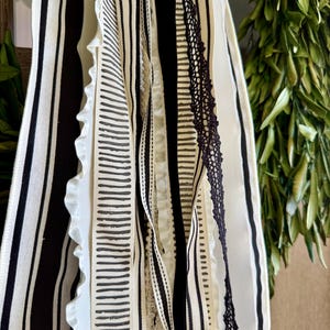 May include: Close-up of decorative ribbons in black and white. The ribbons feature various patterns, including stripes, solid colors, and a lace trim. The ribbons are layered and hanging, with a green leafy background.