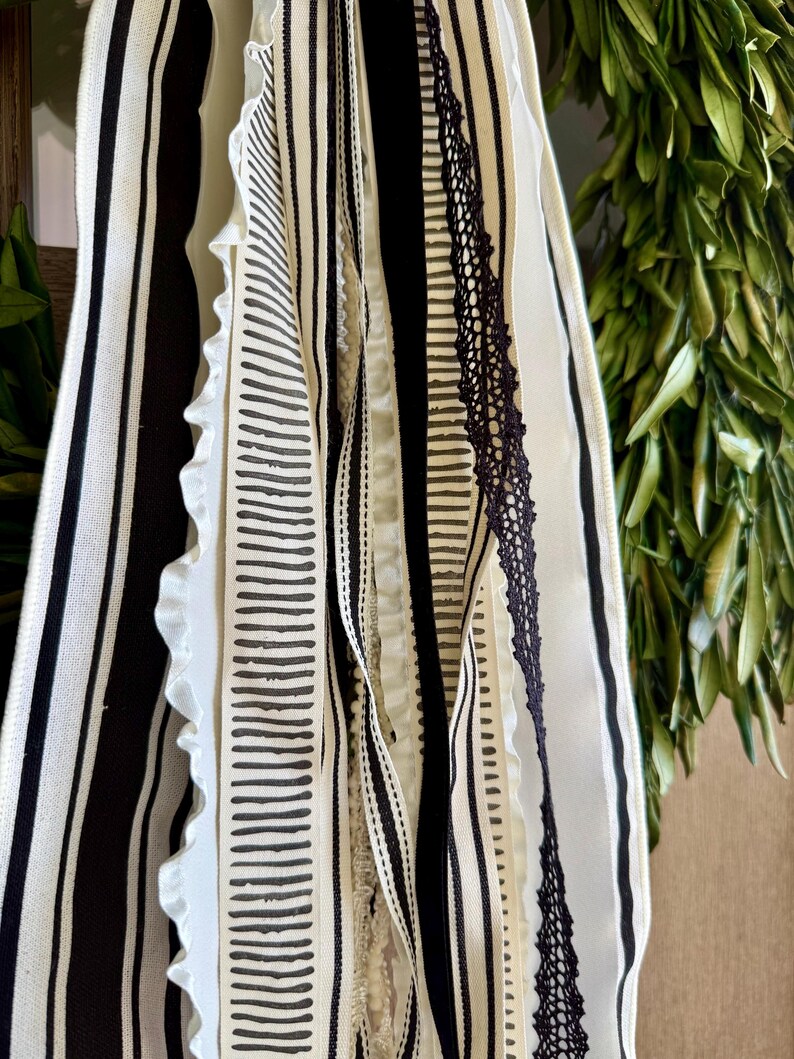 May include: Close-up of decorative ribbons in black and white. The ribbons feature various patterns, including stripes, solid colors, and a lace trim. The ribbons are layered and hanging, with a green leafy background.