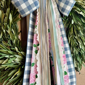 May include: A decorative wreath with a blue and white gingham bow and cascading ribbons. The ribbons feature floral patterns, stripes, and lace, in shades of pink, green, and white. The wreath is made of green leaves.