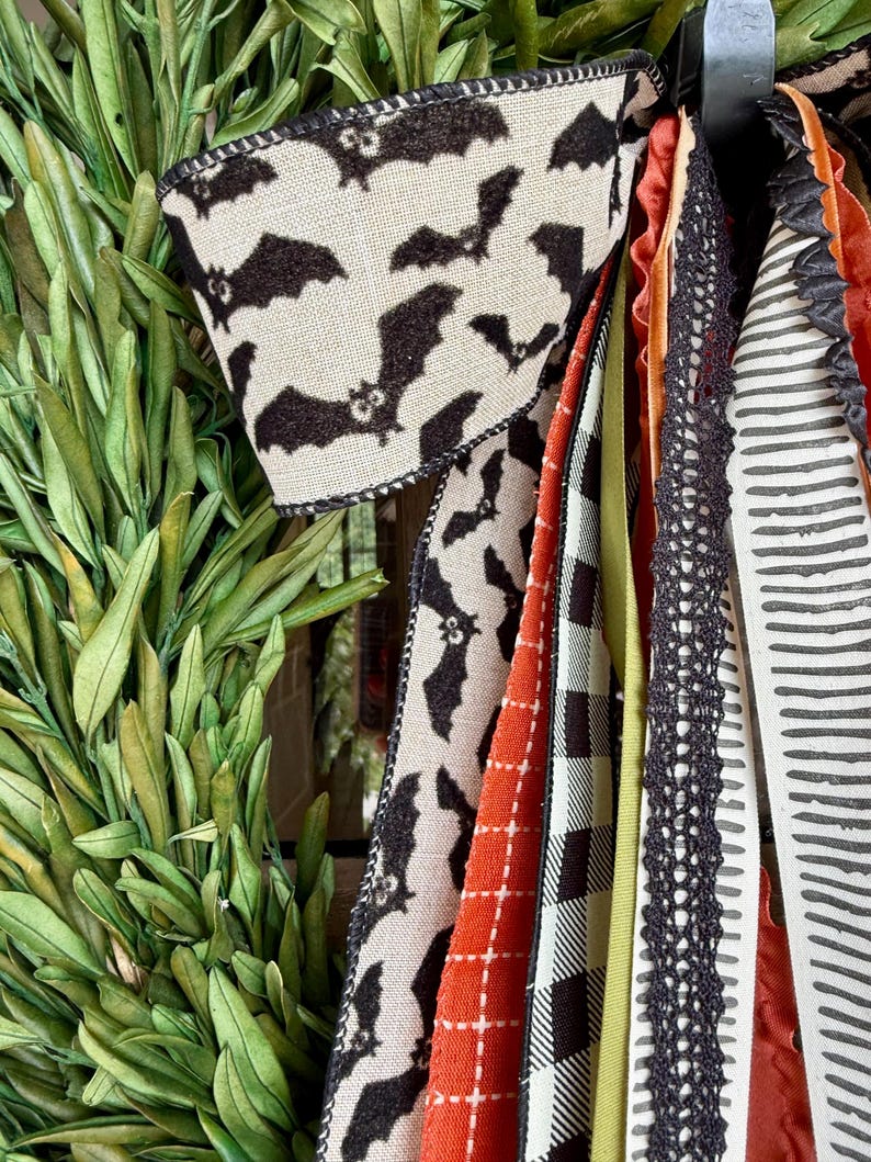 Op de afbeelding: Een Halloween-krans met een verscheidenheid aan decoratieve linten. De linten hebben patronen zoals vleermuizen, ruiten en strepen in kleuren als zwart, oranje en cr&egrave;me. Groen gebladerte is zichtbaar aan de linkerkant van de afbeelding.