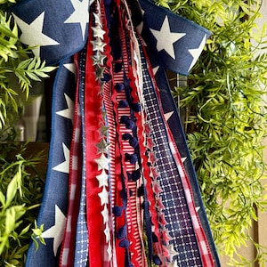 May include: A patriotic wreath with a navy blue bow featuring white stars. Red and white ribbons with star and pom-pom accents cascade down. The wreath is surrounded by green foliage, creating a festive and decorative display.