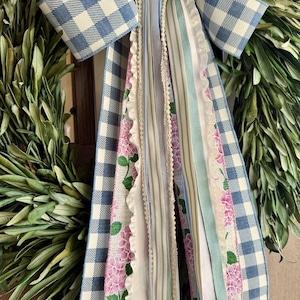 May include: A decorative wreath with a blue and white gingham bow and cascading ribbons. The ribbons feature floral patterns, stripes, and lace in shades of pink, blue, and white. The wreath is adorned with green foliage.