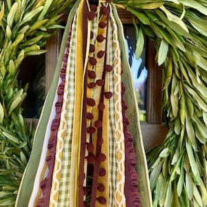 May include: A decorative ribbon bundle featuring multiple textures and colors including olive green, mustard yellow, and deep burgundy. The arrangement includes gingham patterns, velvet-like strips, ruffled edges, and a trim with small brown pom-poms.