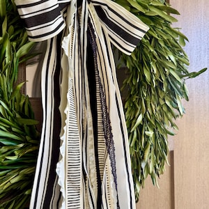 May include: A fresh green olive wreath adorned with a black and white striped bow and cascading ribbons. The ribbons feature various patterns, including stripes and lace, adding texture and visual interest to the natural greenery.