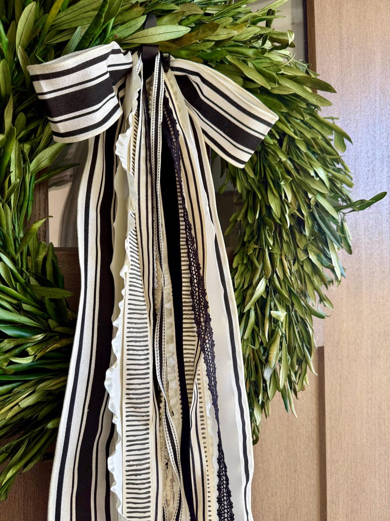 May include: A fresh green olive wreath adorned with a black and white striped bow and cascading ribbons. The ribbons feature various patterns, including stripes and lace, adding texture and visual interest to the natural greenery.