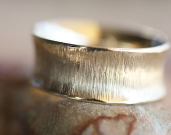 Puede incluir: Primer plano de un anillo dorado con un acabado texturizado y cepillado. El anillo tiene una banda ancha y una forma ligeramente curvada. El anillo está sobre una superficie marrón claro.