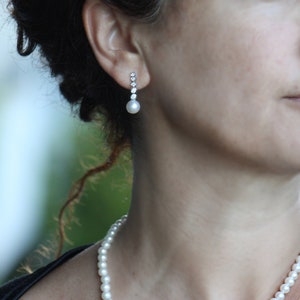 May include: A woman wearing a black top and a pearl necklace. She is wearing a pearl and diamond earring.