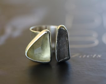 May include: A silver ring with two stones set in it. One stone is a clear, light green triangle, and the other is a dark gray rectangle.