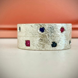May include: A silver ring with a textured surface and a variety of small gemstones in red and blue.