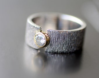 May include: A silver ring with a textured finish and a gold bezel setting holding a clear, round gemstone. The ring has a modern, minimalist design, suitable for everyday wear or special occasions.