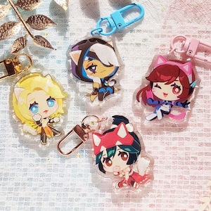 May include: Four colorful character keychains with gold and silver key rings. The characters are drawn in an anime style and feature a pink cat, a blue cat, a yellow cat, and a red cat.