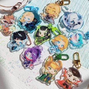 May include: A set of 12 colourful anime character keychains. The keychains are made of clear acrylic and feature various characters with different expressions and poses. The keychains are attached to gold metal rings.