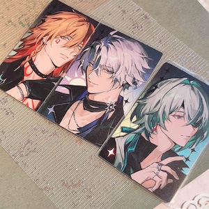 以下が含まれることがあります： アニメスタイルのイラストが描かれた、3枚のキャラクターアートカード。カードには、独特の髪型と服装をした男性の姿が描かれています。カラーパレットは赤、白、青の色合いで、黒のアクセントがあります。各カードには星のデザインがあります。