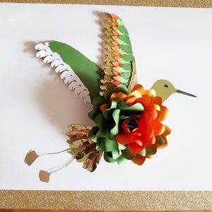 以下が含まれることがあります： 緑、オレンジ、赤の紙の花のアクセントが付いた、紙製のハチドリのクラフト。ハチドリは羽を広げて飛んでいる。