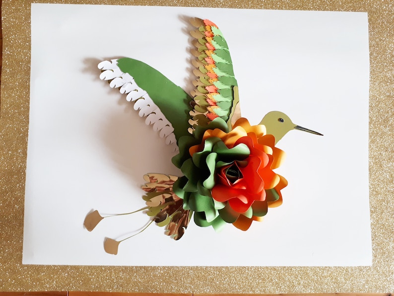 以下が含まれることがあります： 緑、オレンジ、赤の紙の花のアクセントが付いた、紙製のハチドリのクラフト。ハチドリは羽を広げて飛んでいる。