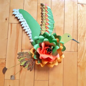 以下が含まれることがあります： 緑、オレンジ、赤の紙の花と、長い嘴と白いアクセントのついた羽根を持つ緑のハチドリ