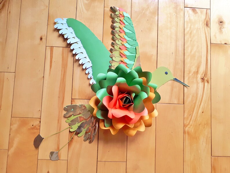 以下が含まれることがあります： 緑、オレンジ、赤の紙の花と、長い嘴と白いアクセントのついた羽根を持つ緑のハチドリ