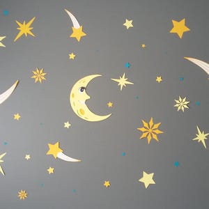 May include: A yellow crescent moon with a face and eyelashes, surrounded by yellow stars and shooting stars on a grey background.