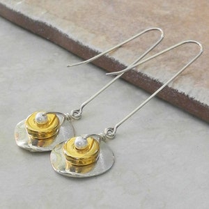 May include: A pair of silver dangle earrings with a gold and white pearl accent. The earrings have a long, thin silver wire that hangs from a small, round silver disc. The disc is topped with a smaller gold disc and a white pearl.